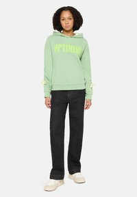 Jasnozielona bluza z kapturem z napisem "OPTIMISM" w żółtej liternictwie, luźny krój, sznurki przy mankietach. W połączeniu z ciemnymi dżinsami i jasnymi trampkami.