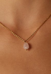 Gouden ketting met een druppelvormige rozenkwarts hangertje, versierd met een kleine gouden kraal, rustend op de huid.