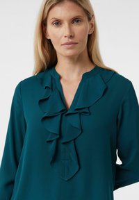 Teal blouse with long sleeves, featuring a ruffled V-neckline and soft, smooth fabric. Simple design without patterns or accents.