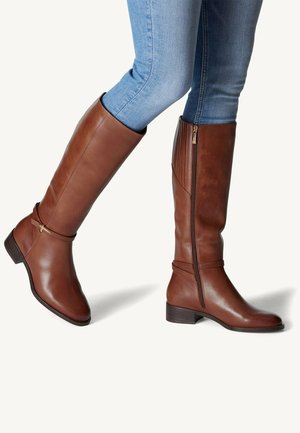Bottes en cuir marron montantes jusqu'au genou avec talons bas et fermetures éclair latérales portées avec un jean skinny bleu clair sur un fond neutre.