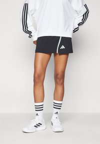 Svart atletisk skjørt med hvit glidelås på siden og logo, parret med hvite sko og stripete sokker. Hvit hettegenser med svarte striper på ermene.