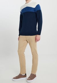 Marineblauer Strickpullover mit strukturierter weiß-blauer obere Partie, kombiniert mit hellbeigen, schmal geschnittenen Hosen und braunen Sneakers.