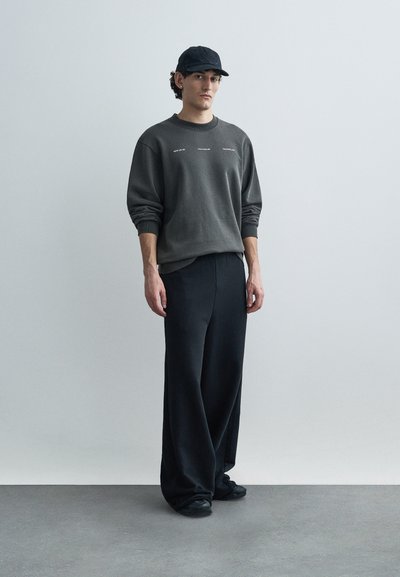 Γκρι φούτερ με στρογγυλή λαιμόκοψη και ριμπ μανσέτες, μαύρο παντελόνι φαρδιάς γραμμής, και μαύρο καπέλο. Ομαλό ύφασμα, άνετη εφαρμογή, μινιμαλιστικός σχεδιασμός.