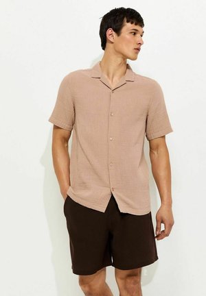 Man draagt een beige kort mouw overhemd met knopen en donkerbruine korte broek, staat met één hand in zijn zak en kijkt naar rechts tegen een witte achtergrond.