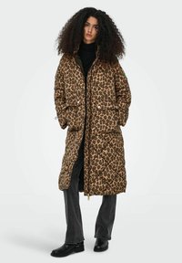 Brązowa pikowana kurtka puchowa w panterkę z zamkiem na przodzie, dwoma przednimi kieszeniami i pikowanym wzorem. Noszona na czarnym topie i szarych spodniach.