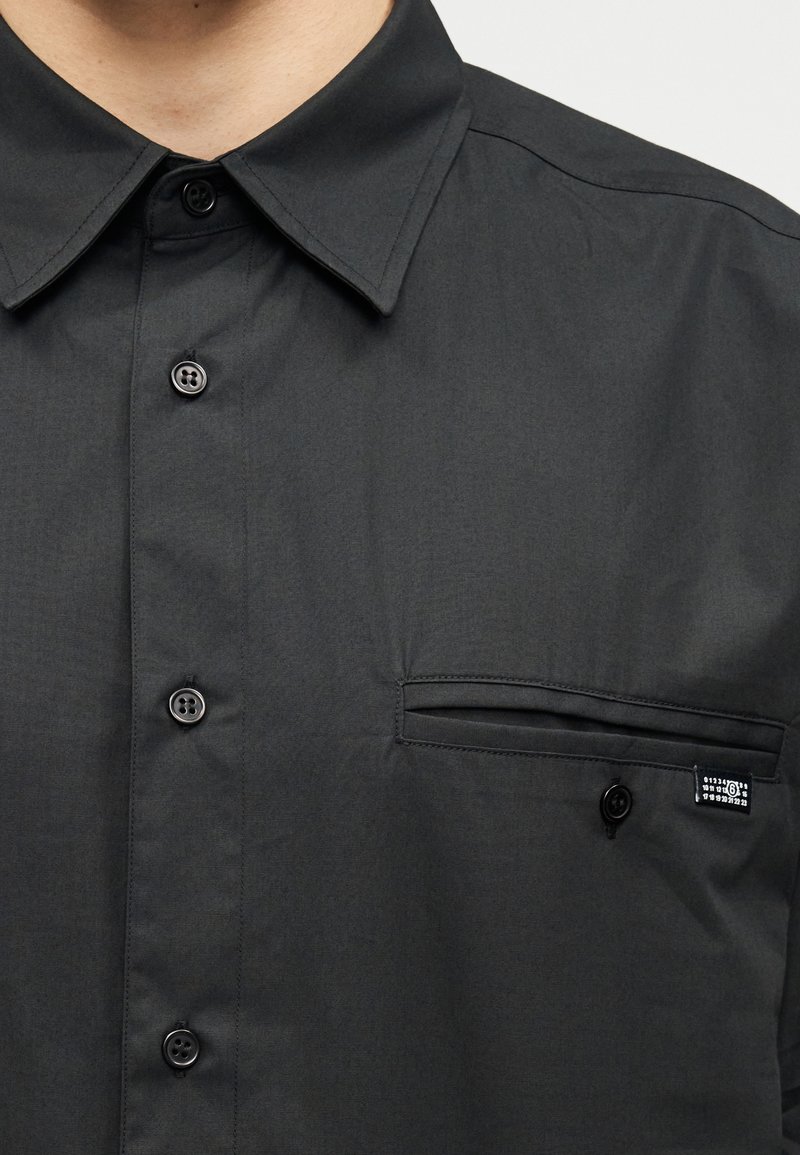 Chemise boutonnée gris foncé avec col pointu, dotée d'une poche poitrine unique, d'une texture subtile et de boutons noirs. Petite étiquette de marque incluse.