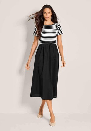 Frau geht nach vorne, trägt einen schwarzen Midirock und ein schwarz-weiß gestreiftes Kurzarmoberteil mit beigen spitzen Schuhen vor neutralem Hintergrund.