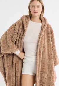 Przewymiarowany, beżowy szlafrok z pluszu z fakturowanym wzorem warkocza, wyposażony w kaptur i otwarty przód. Miękki, puszysty materiał na całej powierzchni.