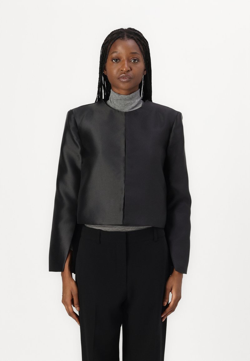 Chaqueta negra corta con un acabado satinado suave, que presenta un cuello alto y mangas largas, combinada con pantalones negros de tiro alto y un suéter de cuello alto gris.