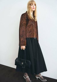Veste marron à pois avec fermeture éclair à l'avant, associée à une longue jupe noire. Des escarpins blancs à lanières et un petit sac à main noir complètent le look.