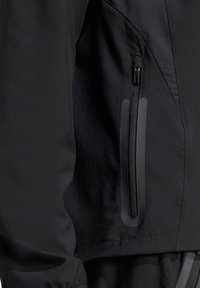 Schwarze Jacke aus einem glatten, leichten Stoff, mit einer Reißverschlusstasche auf der Brust und subtilen strukturierten Akzenten an den Schultern und Ärmeln.