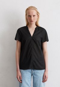 Zwarte blouse met korte mouwen en een V-halslijn, met een geplooide voorkant en een gladde textuur. Gepaard met lichtblauwe spijkerbroek.