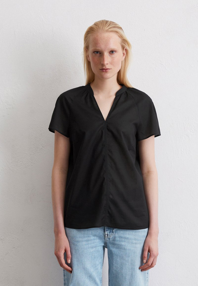 Zwarte blouse met korte mouwen en een V-halslijn, met een geplooide voorkant en een gladde textuur. Gepaard met lichtblauwe spijkerbroek.