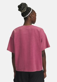 Vrouw met gevlochten haar in een knot, die een witte haarband, een oversized roze T-shirt en zwarte shorts draagt, gezien vanuit de achterkant.