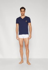 Navy V-neck T-shirt made of cotton, featuring a small emblem on the left chest; paired with white briefs, model standing on a neutral background.