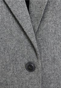 Blazer gris en tissu texturé, avec un bouton noir et des revers pointus. Le motif est composé de fils tissés fins.