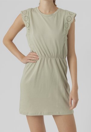 Woman wearing a light green sleeveless dress with eyelet lace ruffle sleeves, elastic waist, standing with one hand on hip.