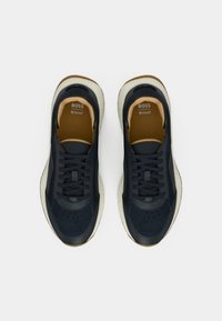 Mustade ja pruunide BOSS-tossude paar valgete tallade ja paeltega, vaadatuna ülalt valgel taustal.