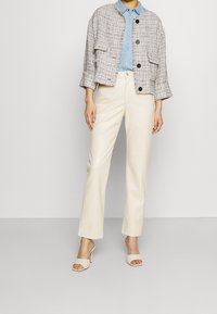 Giacca in tweed con bottoni neri, camicia in denim chiaro, pantaloni crema e scarpe con tacco aperte beige. La giacca ha tasche grandi e una vestibilità ampia.