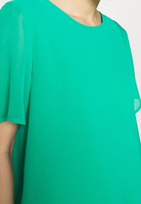 Blusa verde a maniche corte in tessuto leggero; presenta un collo tondo e maniche fluide e semitrasparenti. Tessuto liscio e brillante.