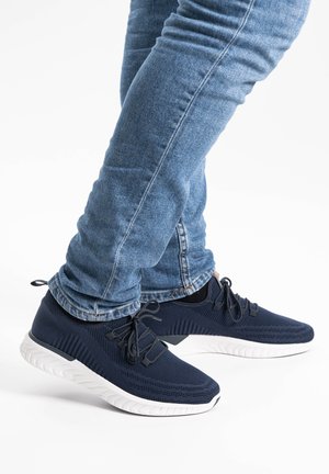 Mørkeblå træningssko med mesh-overdel, hvid sål og kontrasterende sorte detaljer. Båret med lyseblå tætsiddende jeans.