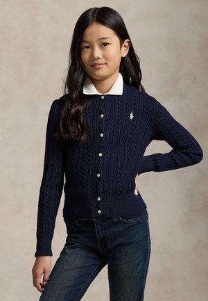 Jeune fille aux longs cheveux foncés portant un cardigan bleu marine en maille torsadée boutonné sur une chemise blanche à col et un jean foncé, debout avec une main sur la hanche.