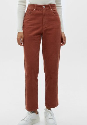 Femme portant un pantalon en velours côtelé marron rouille taille haute et des baskets blanches, debout devant un fond clair uni.