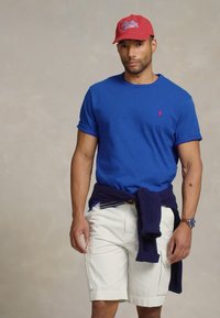 Μπλε βαμβακερό t-shirt, λευκά σορτς, ναυτικό πουλόβερ δεμένο στη μέση και κόκκινο καπέλο με λογότυπο. Ρολόι στον αριστερό καρπό. Απλή, καθημερινή εμφάνιση.