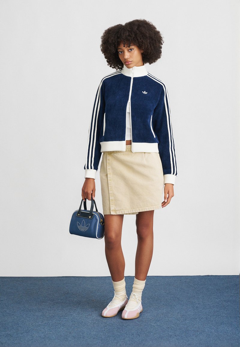 Veste en velours côtelé marine avec des rayures blanches, jupe beige, sac à main bleu et chaussures roses. Les accessoires comprennent des chaussettes crème, créant un look décontracté en superposition.