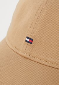 Chaki kepurė, pagaminta iš tekstūruotos medžiagos, su mažu išsiuvinėtu logotipu mėlynos, baltos ir raudonos spalvų šone. Matomi siuvimo detalės.