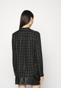 Schwarze karierte Bluse mit langen Ärmeln, aus weichem Stoff und mit einem geknöpften Kragen. Der Rücken verfügt über eine zentrale Naht und ein dezentes Falten-Detail.