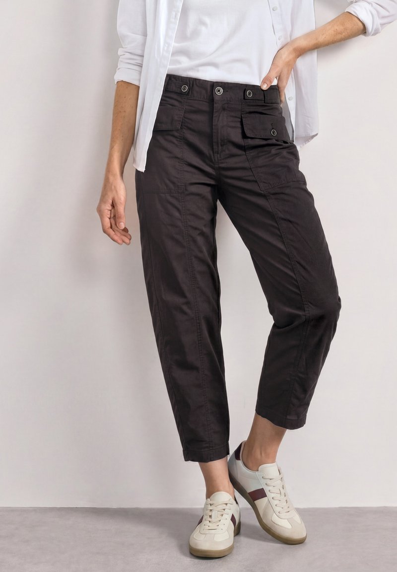 Femme portant un pantalon cargo noir court avec des poches à boutons, des baskets blanches avec des rayures marron, une chemise blanche et des manches retroussées.