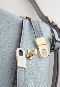 Világoskék bőrtáska arany téglalap alakú nyomógombos zárral és hozzáillő bőr pántokkal, amelyek aranyszínű fémalkatrészekkel vannak rögzítve.