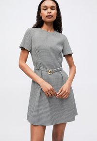 Γυναίκα που φορά ένα κοντομάνικο φόρεμα σε μαύρη και άσπρη καρό (houndstooth) σχέδιο, με ταιριαστό ζώνη που έχει χρυσή αγκράφα, στέκεται μπροστά σε λευκό φόντο.