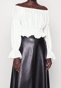 Blusa bianca off-the-shoulder con maniche a sbuffo e orlo arricciato, abbinata a una gonna in pelle nera con una finitura liscia.
