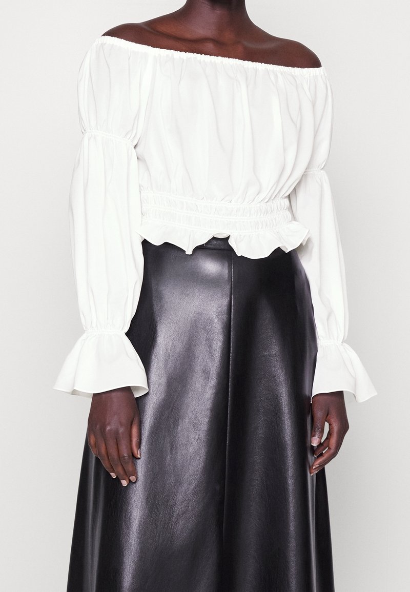 Blusa bianca off-the-shoulder con maniche a sbuffo e orlo arricciato, abbinata a una gonna in pelle nera con una finitura liscia.