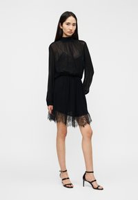 Μαύρο φόρεμα με διαφανή μακριά μανίκια, ψηλό λαιμό και δαντελένιο τελείωμα. Υφασμάτινο σαν σατέν, εφαρμοστή γραμμή και ζώνη στη μέση που δημιουργούν μια κομψή σιλουέτα.