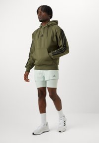 Sweatshirt verde oliva com capuz e riscas pretas nas mangas, combinado com calções curtos de menta claros. Inclui bolso tipo canguru e detalhe do logotipo.
