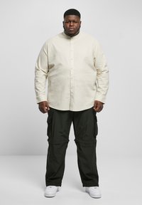 Lichtbeige lange mouwen overhemdd shirt in combinatie met zwarte cargo broek, voorzien van zijzakken en een losse pasvorm. Witte sneakers maken de outfit af.