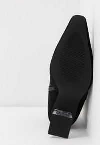 Zapato negro de ante de tacón alto con tacón cuadrado. La suela presenta un patrón texturizado para mayor agarre. Diseño simple sin adornos visibles.