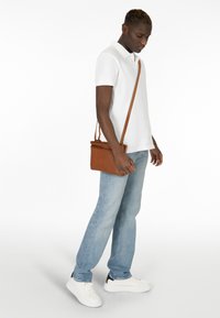 Camicetta polo bianca, jeans azzurri chiari e sneakers bianche. Borsa a tracolla marrone con zip e superficie testurizzata, indossata sulla spalla.