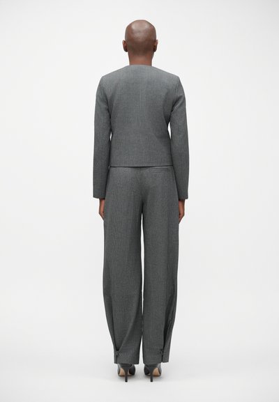 Grijs pak met een gestructureerde afwerking, voorzien van een verkorte blazer en wijde broeken. De broek heeft ritsen bij de enkels en hoge hakken.