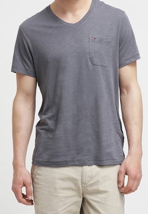 Mand iført en tætsiddende grå V-hals T-shirt med en venstre brystlomme og beige bukser mod en neutral baggrund.