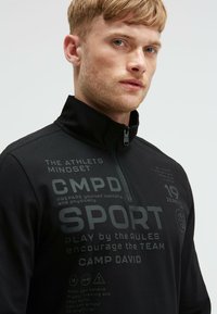 Schwarzer Sportpullover mit Viertelreißverschluss, der mit strukturiertem Material, reflektierenden Grafiken und auffälligem Drucktext, einschließlich Markenname und motivierenden Phrasen, ausgestattet ist.