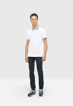 Polo bianco con colletto grigio, due bottoni e maniche corte. Abbinato a pantaloni scuri e sneakers blu navy, in piedi contro uno sfondo neutro.