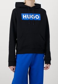Sudadera negra con el logo azul de "HUGO", cordones ajustables, bolsillo frontal y puños acanalados, combinada con pantalones de chándal azules.