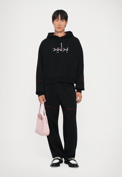 Czarna bluza z kapturem z czerwonymi szwami, w oversize'owym kroju, z graficznym logo. Czarne spodnie, czarne buty z białymi akcentami oraz różowa torebka.