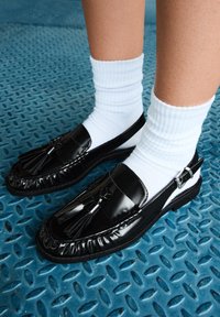Schwarze Lacklederloafer mit Quasten, runde Zehenform und flache Sohle, getragen mit weißen gerippten Socken auf einer strukturierten blauen Oberfläche.