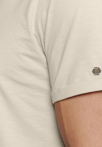  Lichtbeige shirt met korte mouwen van een gestructureerde stof. Heeft een opgerolde manchette en een klein hexagonaal embleem met horizontale lijnen op de mouw.