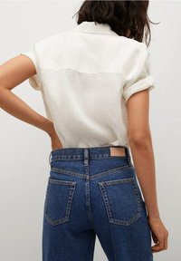 Camisa de lino blanca de manga corta con puños enrollados, combinada con unos vaqueros de mezclilla azul oscuro de cintura alta que cuentan con bolsillos traseros y una etiqueta de cuero.